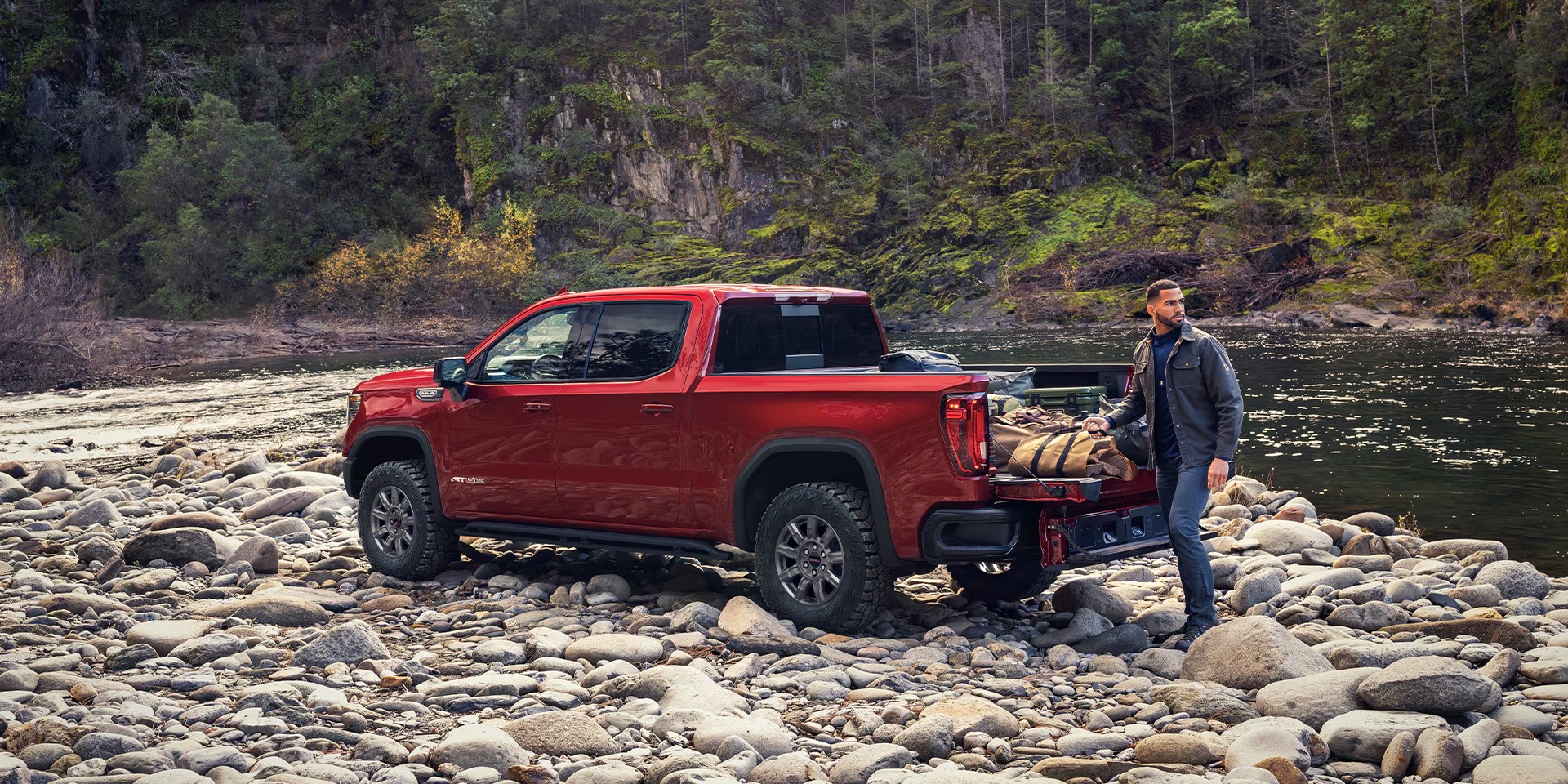 Red 2026 GMC Sierra 1500 AT4X parked on a river rock bed with the tailgate lowered while a person loads gear into the bed