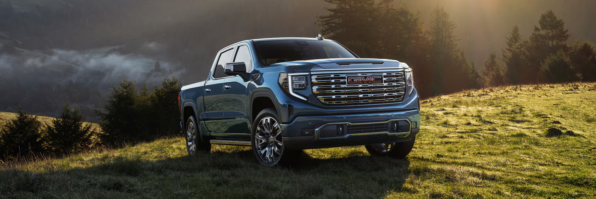 2026 GMC Sierra 1500 Denali parked on a grass hillside at sunrise with mist in the valley behind it