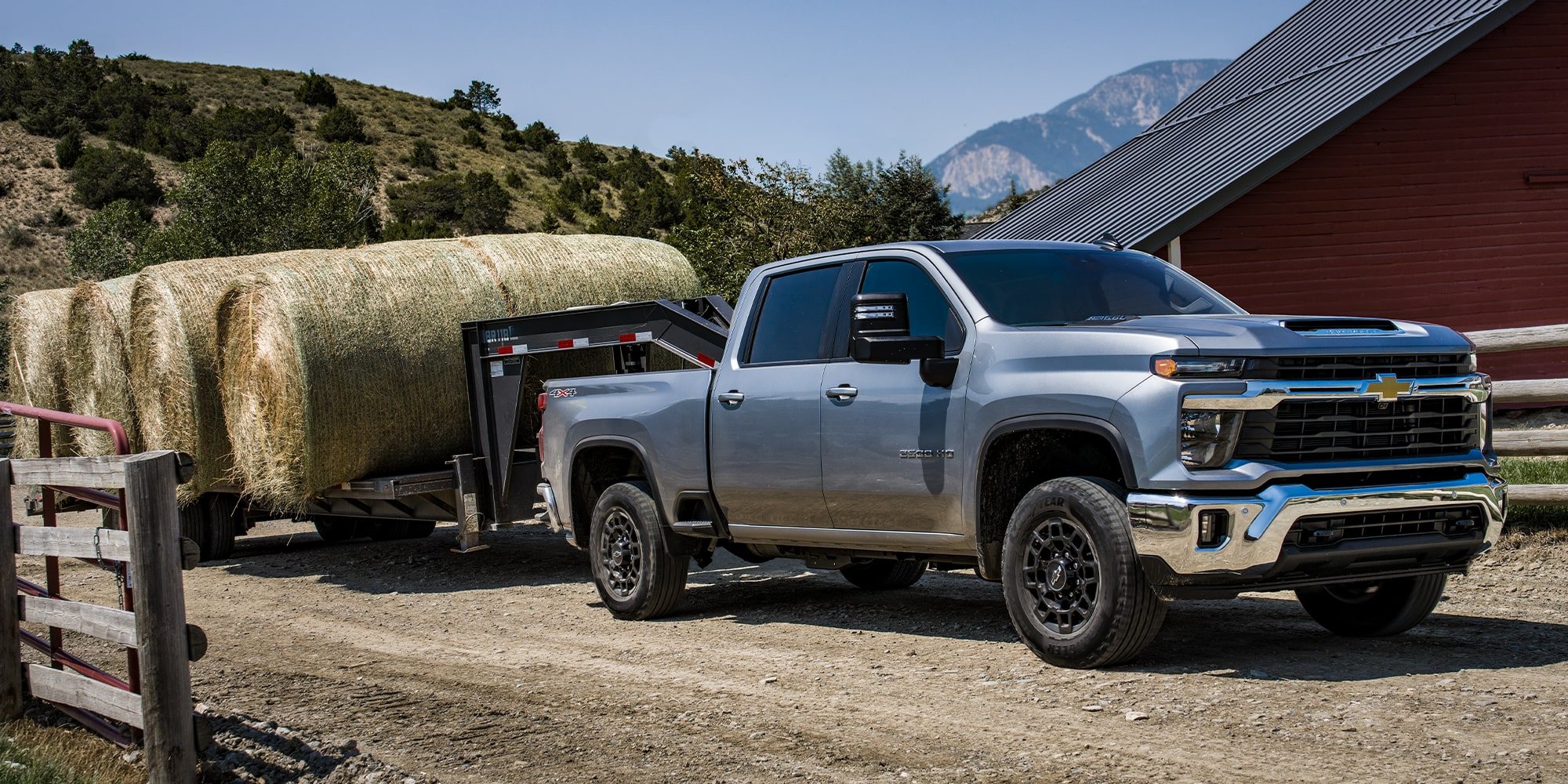 Silver Chevrolet Silverado 2500 HD towing a gooseneck trailer loaded with round hay bales past a red barn