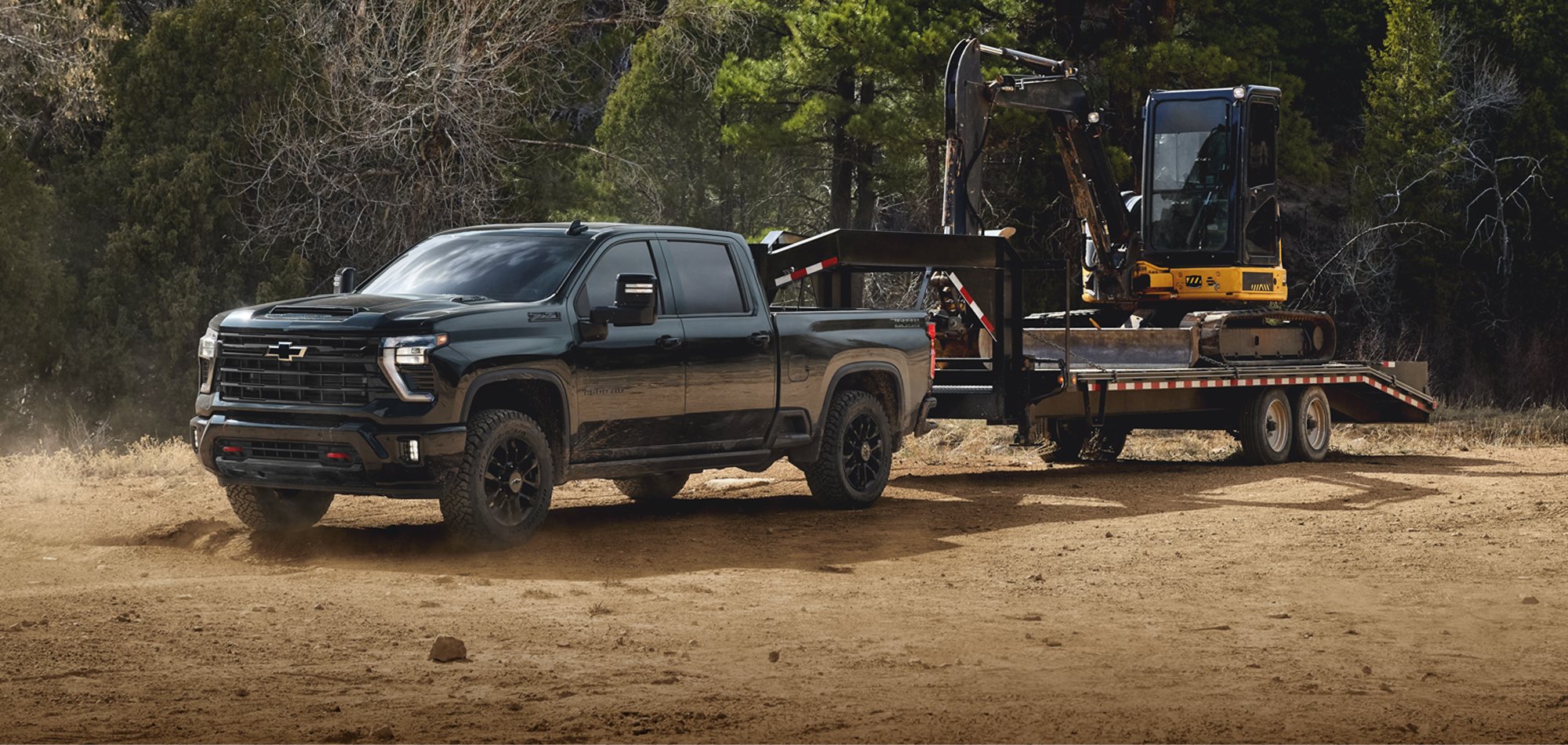 Black Chevrolet Silverado HD crew cab towing a mini excavator on a gooseneck flatbed trailer across a dirt lot, with pine trees behind it