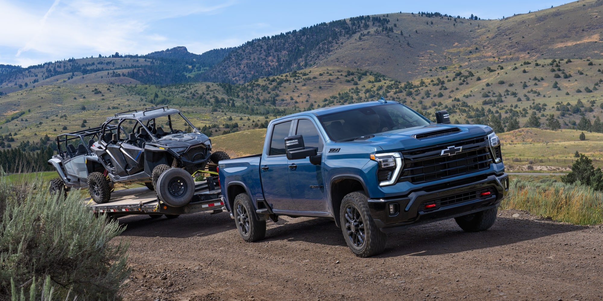 Blue Chevrolet Silverado HD towing a side-by-side on a trailer along a mountain dirt road