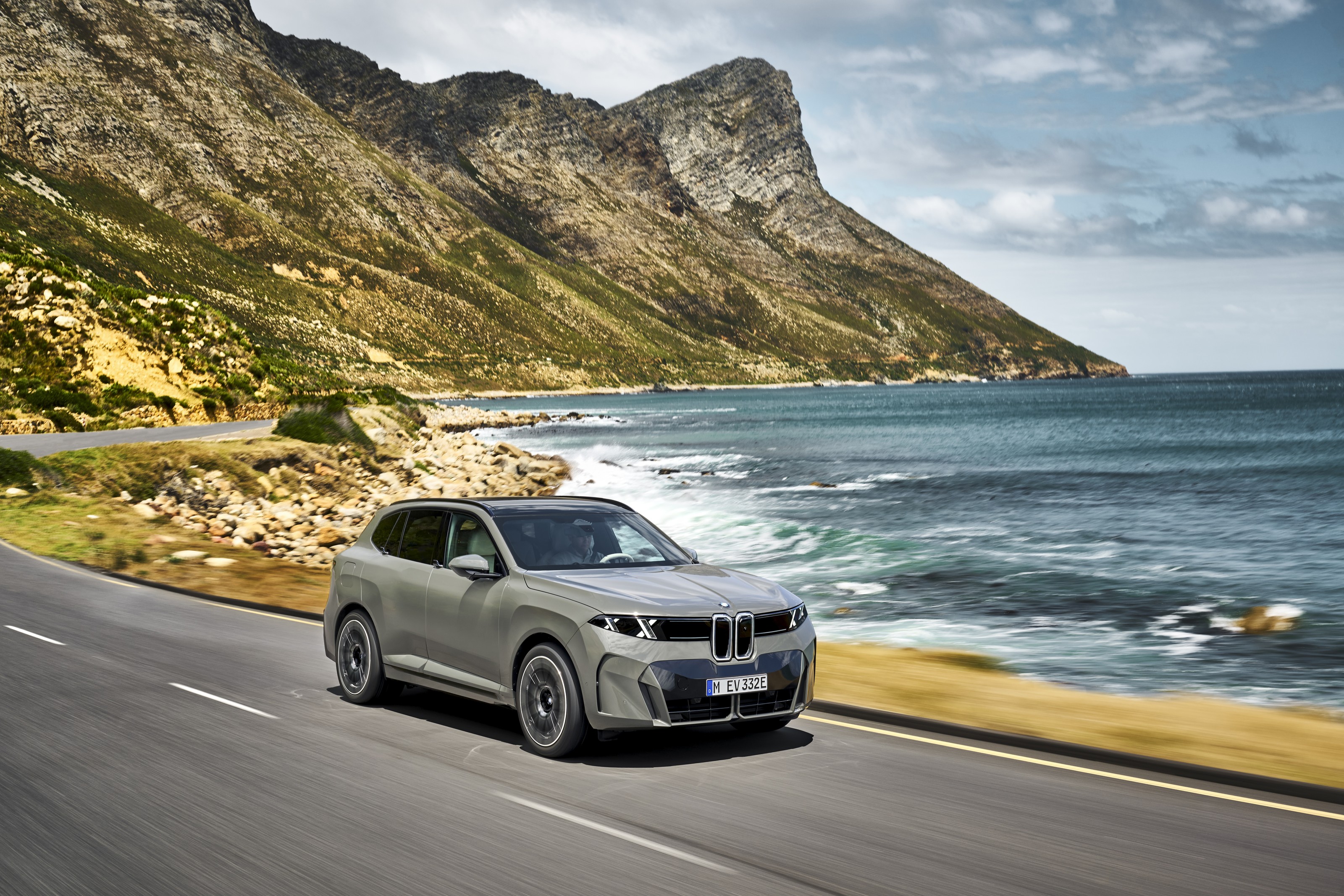 Light gray BMW iX5 driving along a coastal road with a mountain and the sea behind it