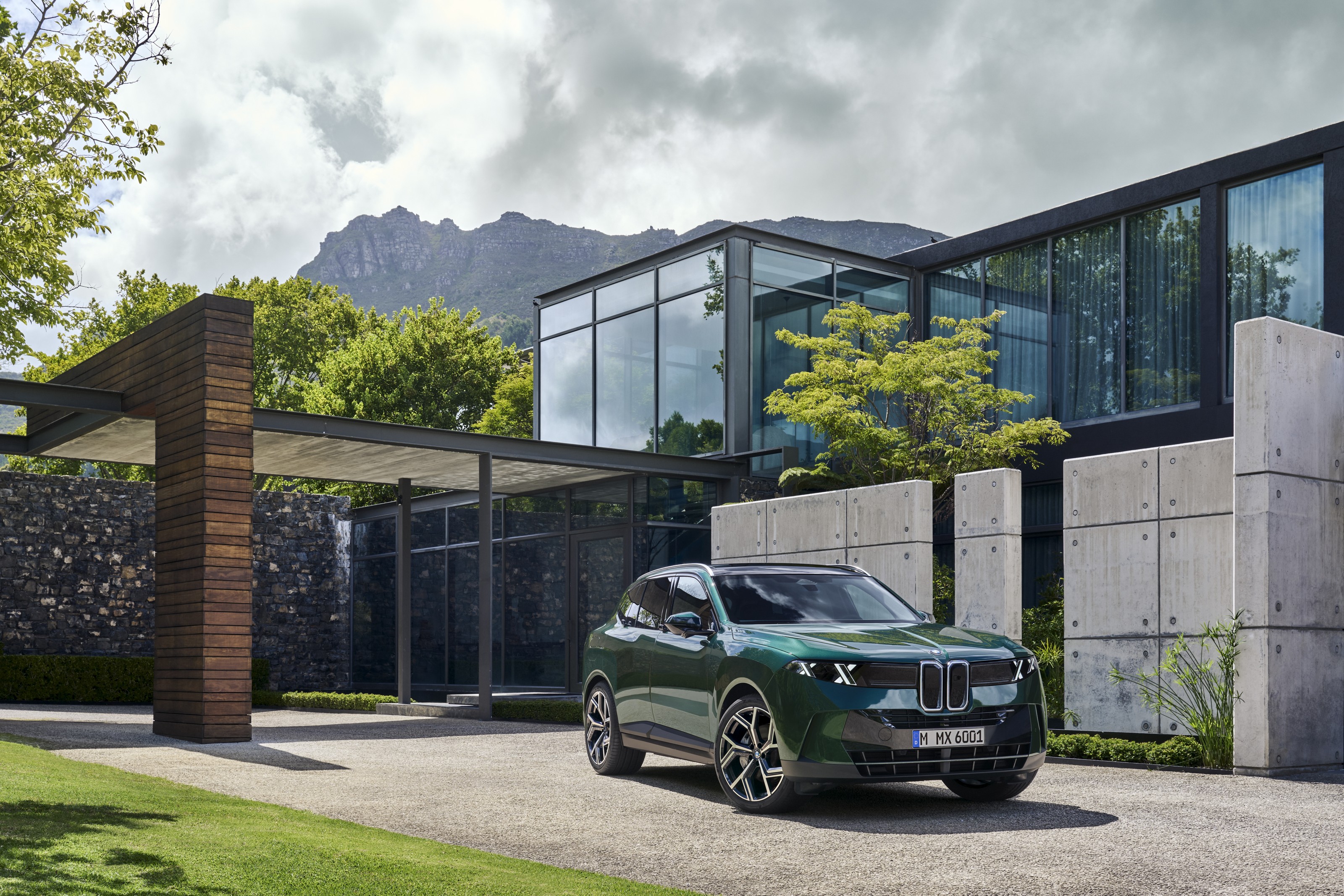 Dark green 2027 BMW X5 parked in the driveway of a modern glass and concrete house, hills behind