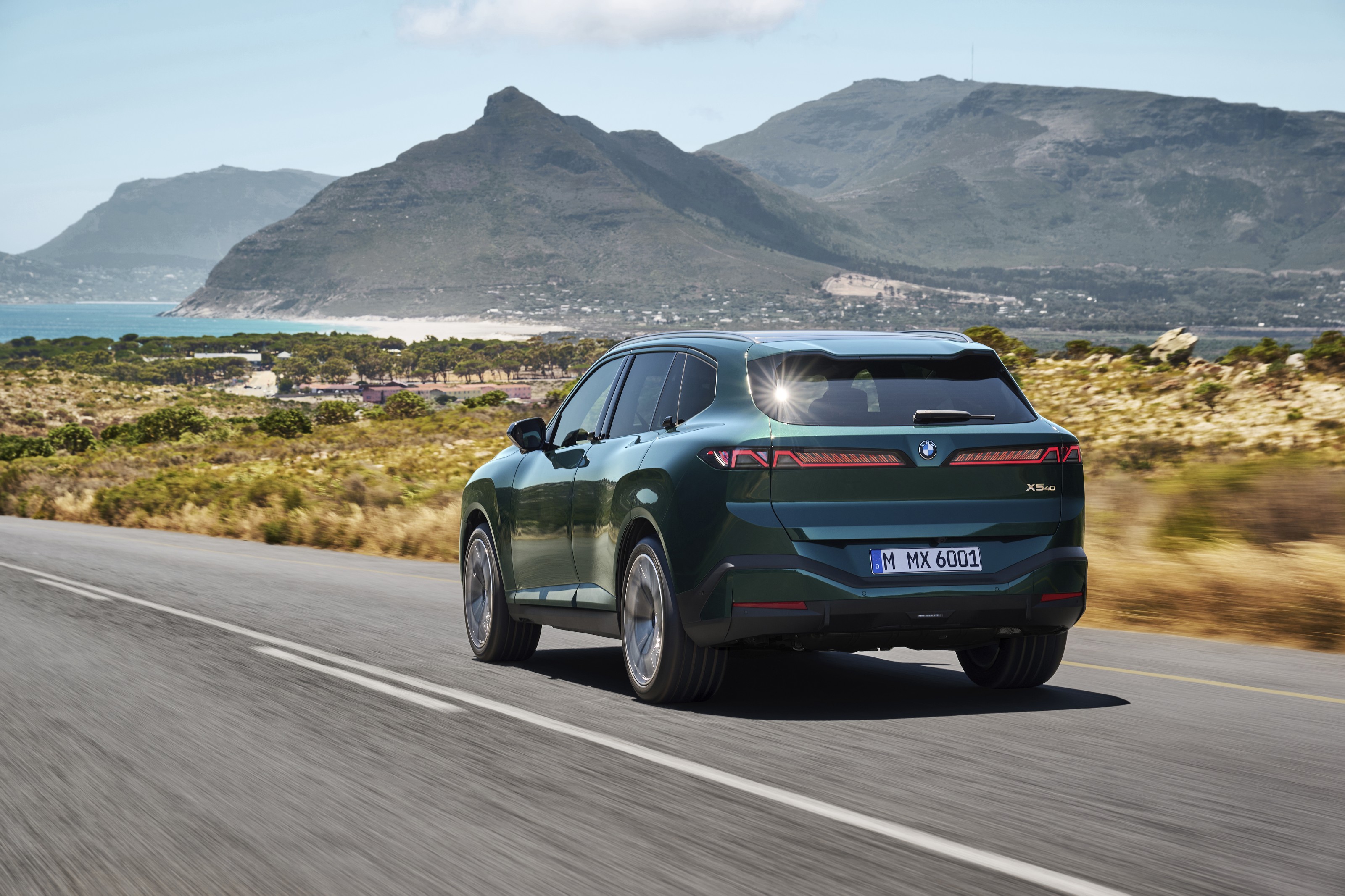 Dark green 2027 BMW X5 driving away along a road through coastal scrubland, with mountains and the sea behind