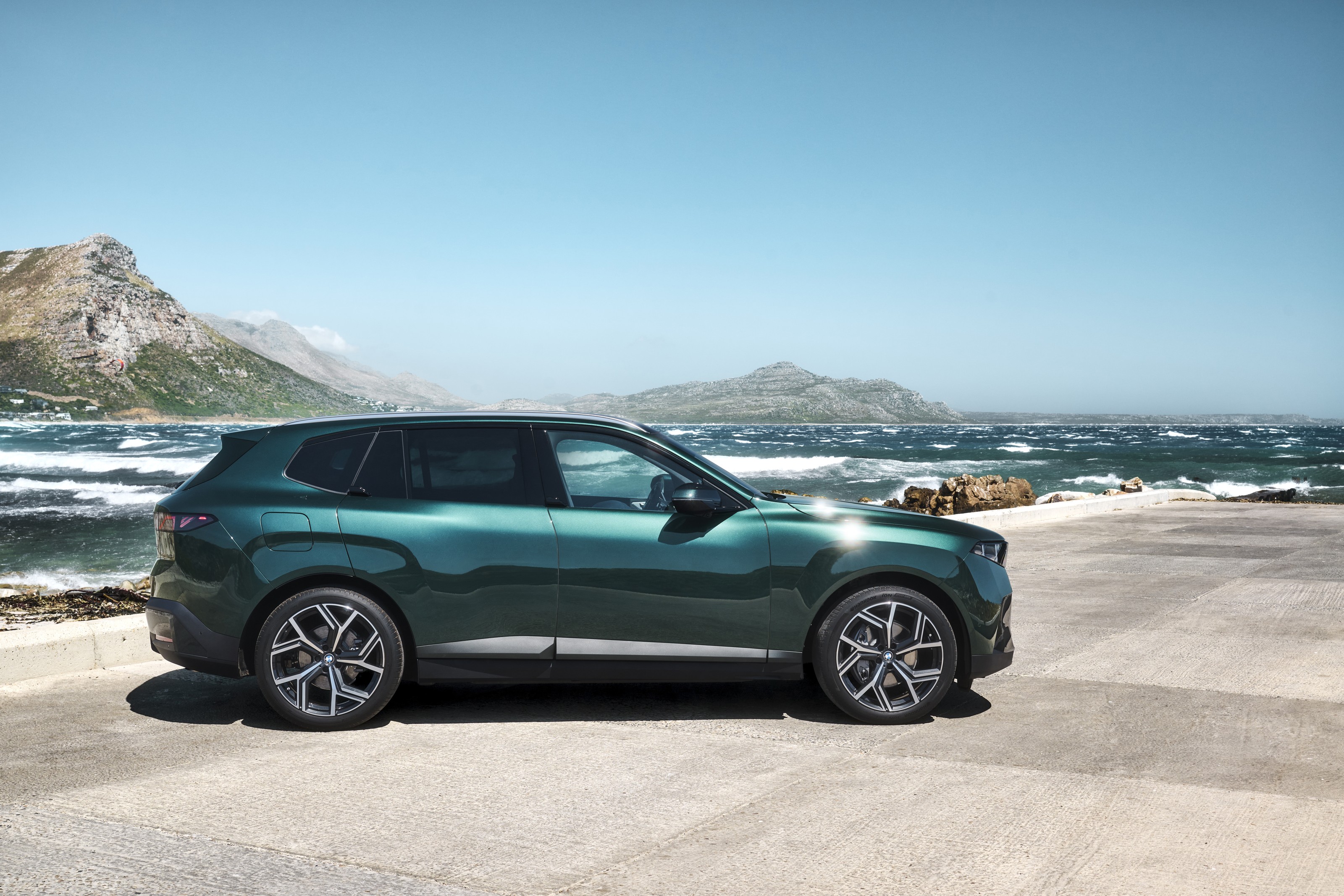 Side profile of a dark green 2027 BMW X5 parked on a seaside lot, with surf and a headland behind it