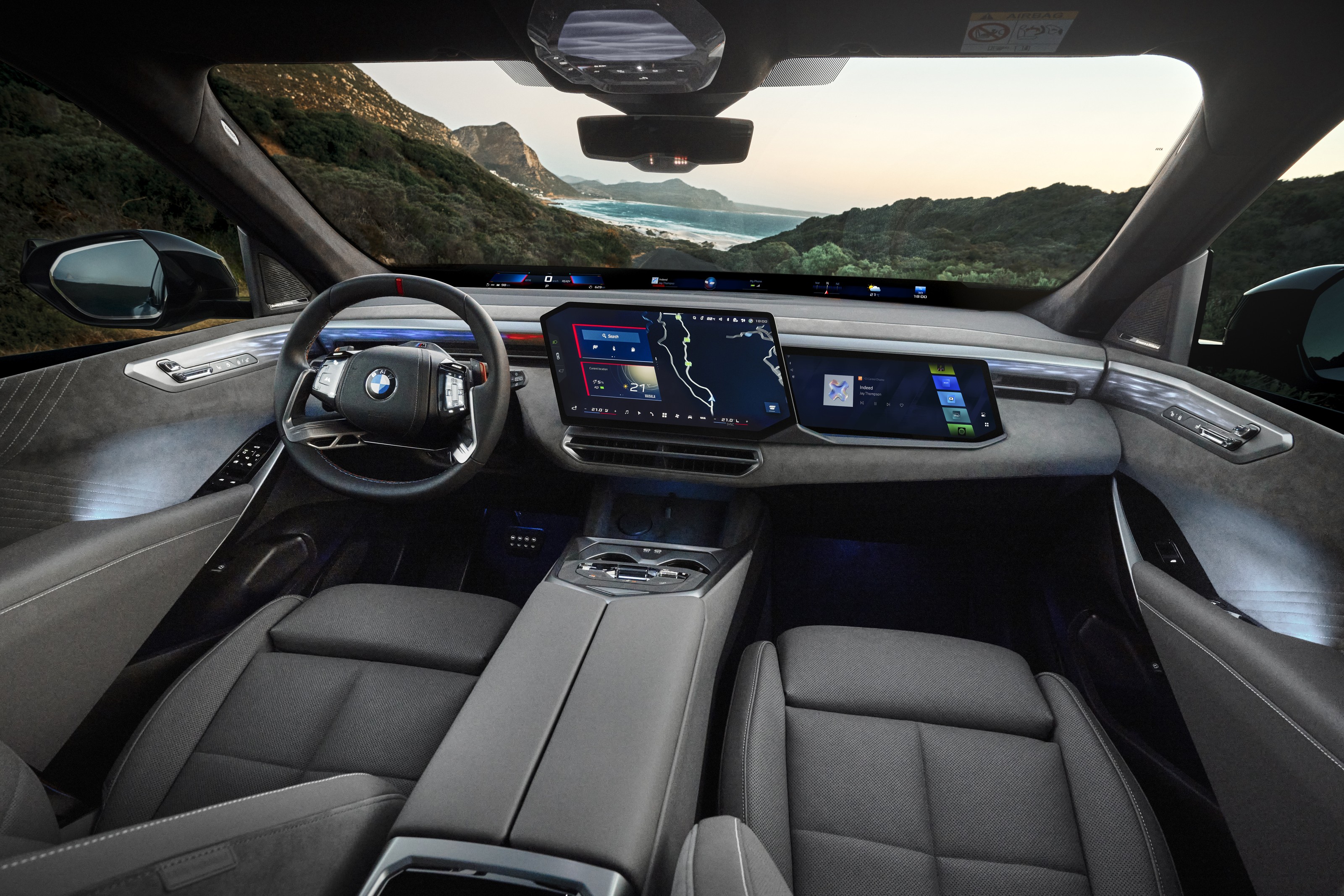 Interior of the new BMW X5 seen from the rear seat: steering wheel, curved central touchscreen and a display band along the windshield base, with a coastal view outside